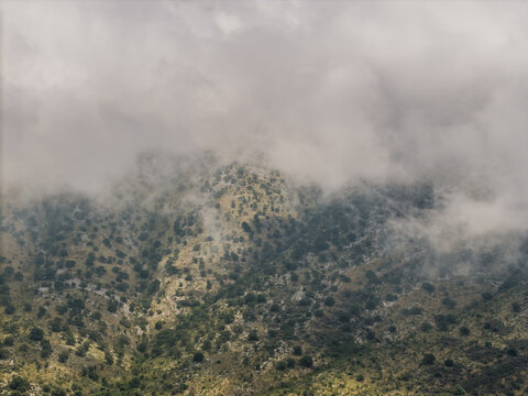 Aerial view of the rugged, cloud-covered mountains near Kroreza Beach, featuring rocky slopes, scattered green vegetation, and thick mist rolling over the terrain in Sarande, Vlore County, Albania.