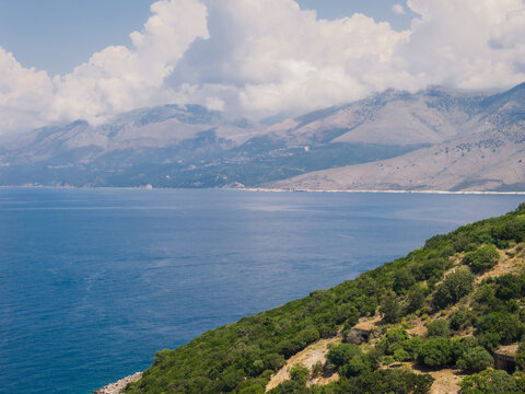 Aerial view of Kepi i Qefalit featuring lush green hills sloping down to the deep blue Ionian Sea under a cloudy sky in Sarande, Albania.