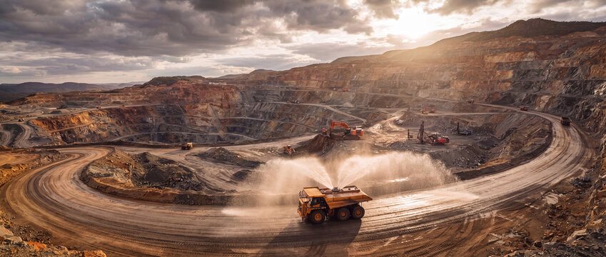 Open pit mine water truck spraying dust along a terraced quarry road with heavy excavators under dramatic sunset clouds, representing industrial mineral extraction operations