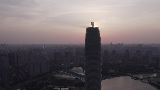 A tall skyscraper stands prominently in a sprawling urban cityscape during dusky hours, with adjacent water bodies and surrounding buildings under a muted sky.