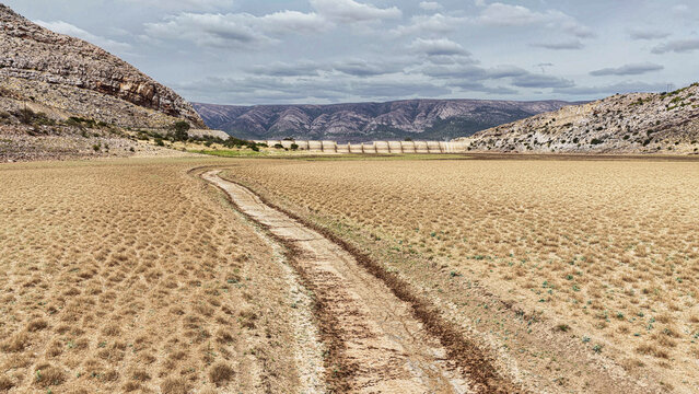 Aerial view of the Karoo landscape with a dry earth path leading towards a distant dam wall, set against rocky hills under a cloudy sky in Beaufort West, Western Cape, South Africa.