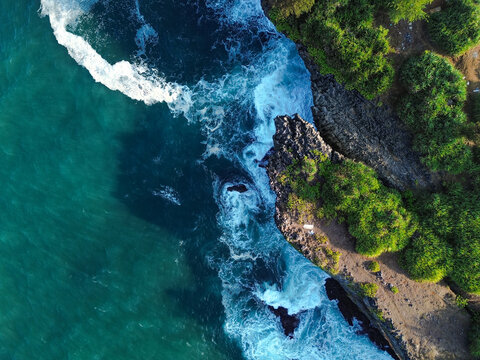 Aerial view of Baron Beach with turquoise waves crashing against rugged cliffs and lush green vegetation in Yogyakarta, Special Region of Yogyakarta, Indonesia.