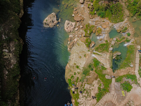 Aerial view of Sri Gethuk Waterfall with turquoise river water, rocky banks, lush greenery, and people swimming in the river in Yogyakarta, Special Region of Yog, Indonesia.