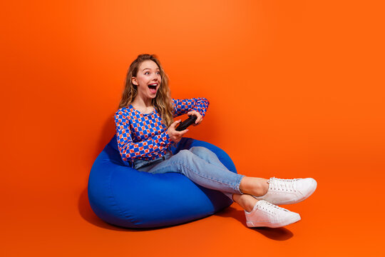 Excited young woman in fashionable attire holding a remote control while relaxing in casual style on an orange background