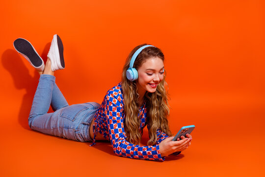Charming woman in casual checkered top wearing headphones enjoying leisure time with smartphone on bright orange background