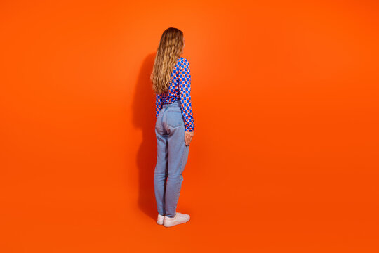 Young blonde woman in checkered top and casual jeans posing against vibrant orange background in studio