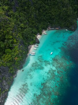 Aerial view of Coron featuring turquoise waters, coral reefs, limestone cliffs, and traditional outrigger boats moored along the sandy beach in Coron, Mimaropa, Philippines.