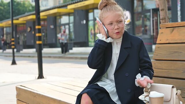 A little girl in a business suit is talking on her smartphone while sitting on a wooden bench outside.
