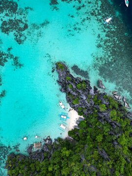 Aerial view of Coron with turquoise water, limestone cliffs, lush vegetation, and traditional outrigger boats anchored along the white sand beach in Coron, Mimaropa, Philippines.