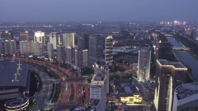 Aerial panorama of a modern urban area at dusk, featuring high-rise buildings, a river, and illuminated roads. 