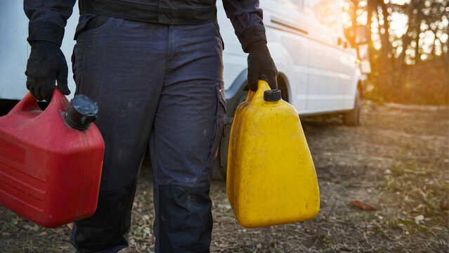 Worker carrying red and yellow plastic fuel canisters near white van outdoors close up
