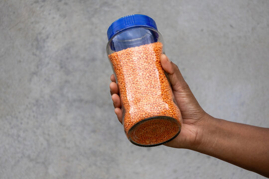 Hand holding transparent glass jar filled with red split lentils against rustic wall. Raw masoor dal stored in container for kitchen household pantry.