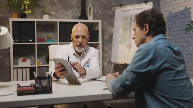 Doctor consults male patient, showing health check-up results on digital tablet