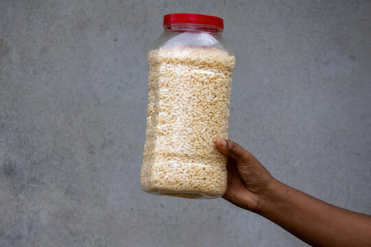 Person holding plastic container filled with crispy puffed rice against rustic grey wall background. Traditional South Asian muri stored for daily tea snack.