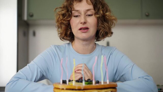 Woman with curly hair blowing out candles on birthday cake. Light blue top, neutral kitchen background, 25 fps