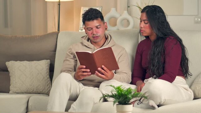 Husband reading the Bible aloud to his wife on a sofa at home