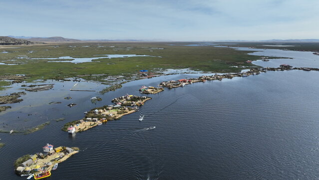 Aerial view of the Uros floating islands on Lake Titicaca featuring traditional reed houses, small boats navigating the blue water, and vast wetlands in Urus, Peru.