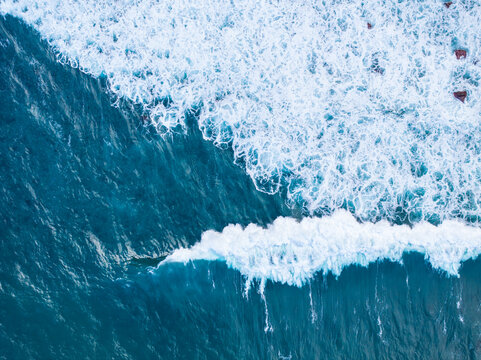 Aerial view of ocean waves crashing against the rocky coastline with deep blue water and white sea foam, Madeira, Portugal.