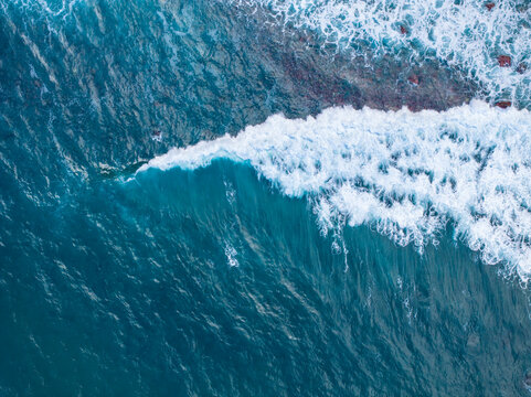 Aerial view of crashing ocean waves with white foam and deep blue water along the rocky coastline of Madeira, Madeira, Portugal.