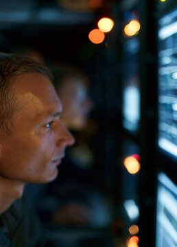 Close-up of a man's face in front of multiple computer monitors. the man appears to be in a control room or a server room, as there are multiple rows of monitors on the right side of the image.