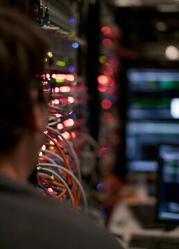 Person's back view of a server room with multiple computer monitors and cables. the person is sitting in front of a rack of servers and appears to be working on a computer.