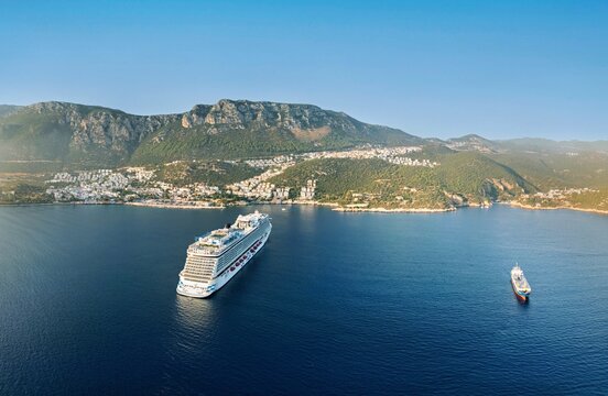 Aerial view of a large cruise ship anchored in the Mediterranean Sea near the coastal town of Kas, with lush green hills and white buildings in the background, Kas, Antalya, Turkey.