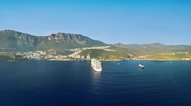 Aerial view of a large cruise ship anchored in the deep blue waters of a bay near the coastal town of Kas, with green mountains in the background, Kas, Antalya, Turkey.