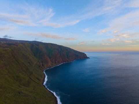 Aerial view of the dramatic coastline of Madeira, featuring steep green cliffs plunging into the deep blue Atlantic Ocean under a soft sky at sunset Madeira, Madeira, Portugal.