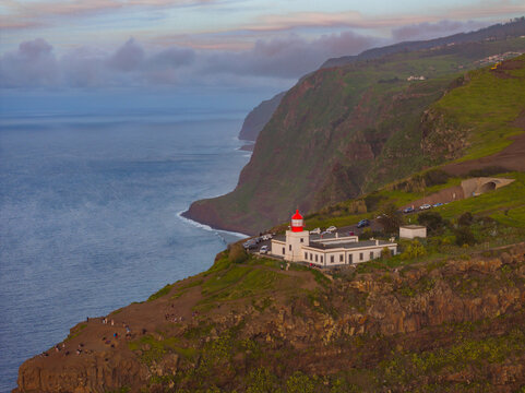 Aerial view of the Ponta do Pargo Lighthouse on a dramatic cliff edge overlooking the Atlantic Ocean at sunset, with green slopes and a coastal road in Ponta do Pargo, Madeira, Portugal.