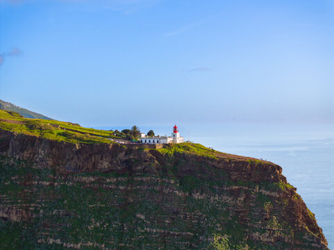 Aerial view of Ponta do Pargo Lighthouse perched on a high cliff overlooking the Atlantic Ocean, surrounded by green slopes and clear blue sky in Madeira, Madeira, Portugal.