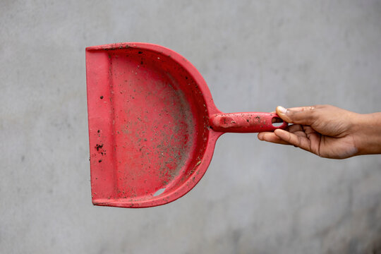 Person holding dirty red plastic dustpan scoop used for gathering floor dust. Ideal for home maintenance work, sanitation tasks, and housekeeping.