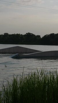 Various ramps and kickers for wakeboarding float on the calm surface of a lake surrounded by trees under an overcast sky. Concept of extreme sports infrastructure and summer recreation.