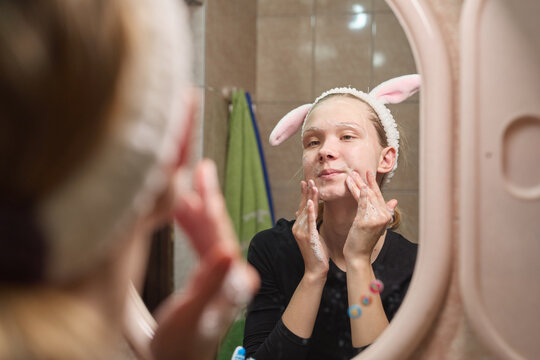 A young girl washes her face in front of the mirror. She applies foam to her face.