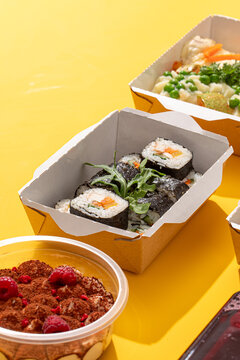 Takeaway meal box with nori sushi rolls dessert with raspberries and healthy dishes on bright yellow background