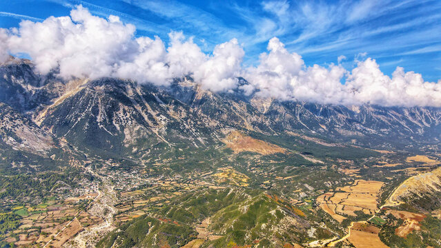 Aerial view of the rugged Llogara mountains with clouds clinging to the peaks, green slopes, and agricultural fields in the valley below in Radhime, Qarku i Vlores, Albania.