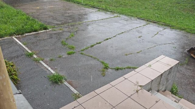 High angle static video of heavy rain falling on old cracked asphalt with weeds and a tiled staircase.