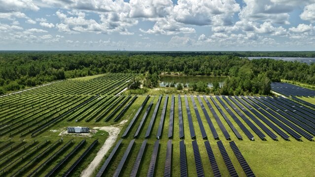 Aerial view of Magnolia Solar Farm with rows of photovoltaic panels, green grass, surrounding forest, and a small pond under a blue sky in Green Cove Springs, Florida, United States.