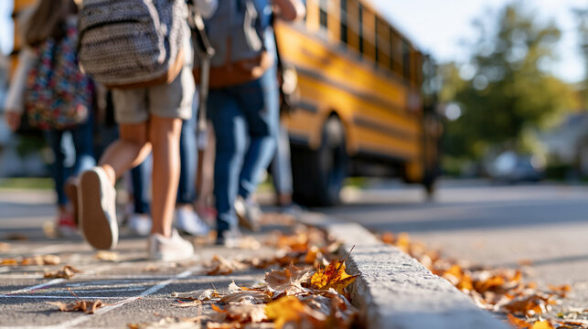 Cropped children with colorful patterned backpacks boarding a bright yellow school bus on a sunny autumn day crunchy fallen leaves scattered along the curb and a chalk hopscotch grid