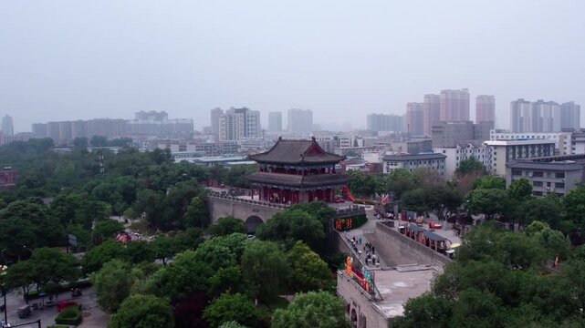 A historic traditional Chinese tower stands among dense green trees, with modern city buildings in the background.
