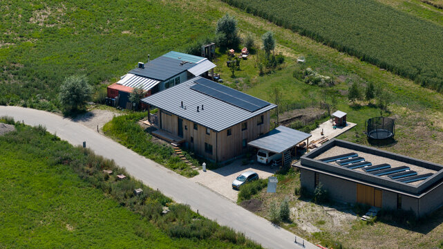 Aerial view of a modern wooden house with solar panels and a separate brick structure situated in a rural landscape with green fields and a road in Almere, Flevoland, Netherlands.