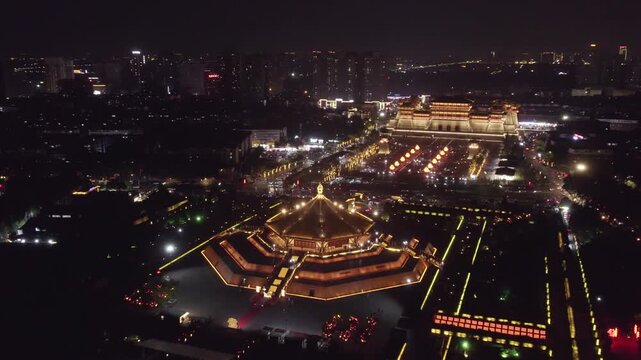 Aerial night shot of an illuminated traditional Chinese architectural complex in an urban area, showcasing historic structures at night.