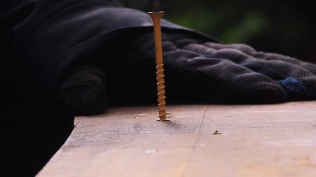 Close-up angled view of a power drill driving a screw into a wooden board, with wood shavings collecting around the screw as it penetrates the surface.