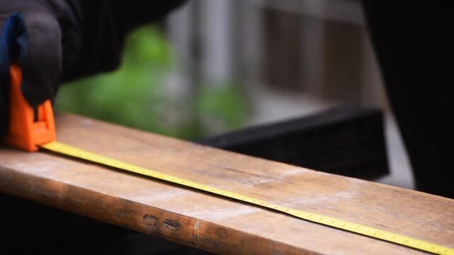 Close-up angled view of a person using a tape measure to measure and mark a wooden board lengthwise, illustrating precision carpentry, woodworking, and DIY construction.
