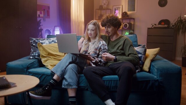 Young couple sit on couch in living room. Two young people are lively share thoughts and look at screen. Female is holding laptop and smile. Guy is gesturing and hugging shoulders of girl.