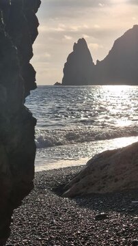 Black sea landscape showing rocky mountains and gentle surf at sunset on a pebble beach. Static eye level shot.