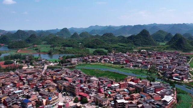 An aerial view of a rural settlement nestled among karst mountains and water bodies under a clear sky.