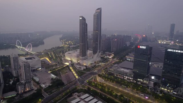 A twilight aerial view of modern twin skyscrapers beside a river in an urban commercial district with lit roads and buildings.