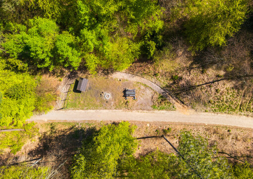 Aerial view of a clearing in Lasy Oliwskie with a wooden shelter, fire pit, and bench along a dirt path in the forest in Gdansk, Pomeranian Voivodeship, Poland.