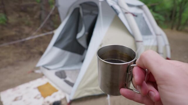 A person holds a stainless steel metal mug filled with dark coffee at a peaceful forest campsite. A modern inflatable camping tent sits blurred in the background among green trees.