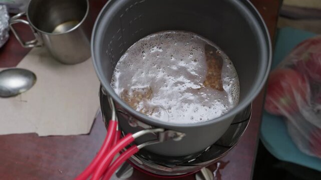 Buckwheat groats boil inside a compact camping pot placed over a portable burner stove. White foam rises as water boils during outdoor food preparation on a trekking adventure.